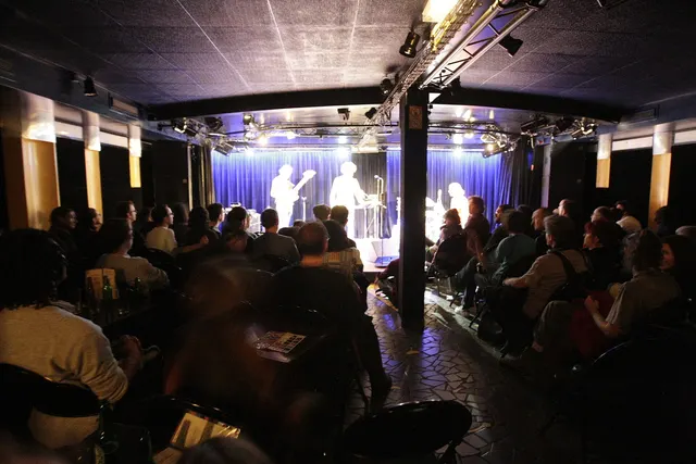Audience seated in front of the lit stage of a small concert hall